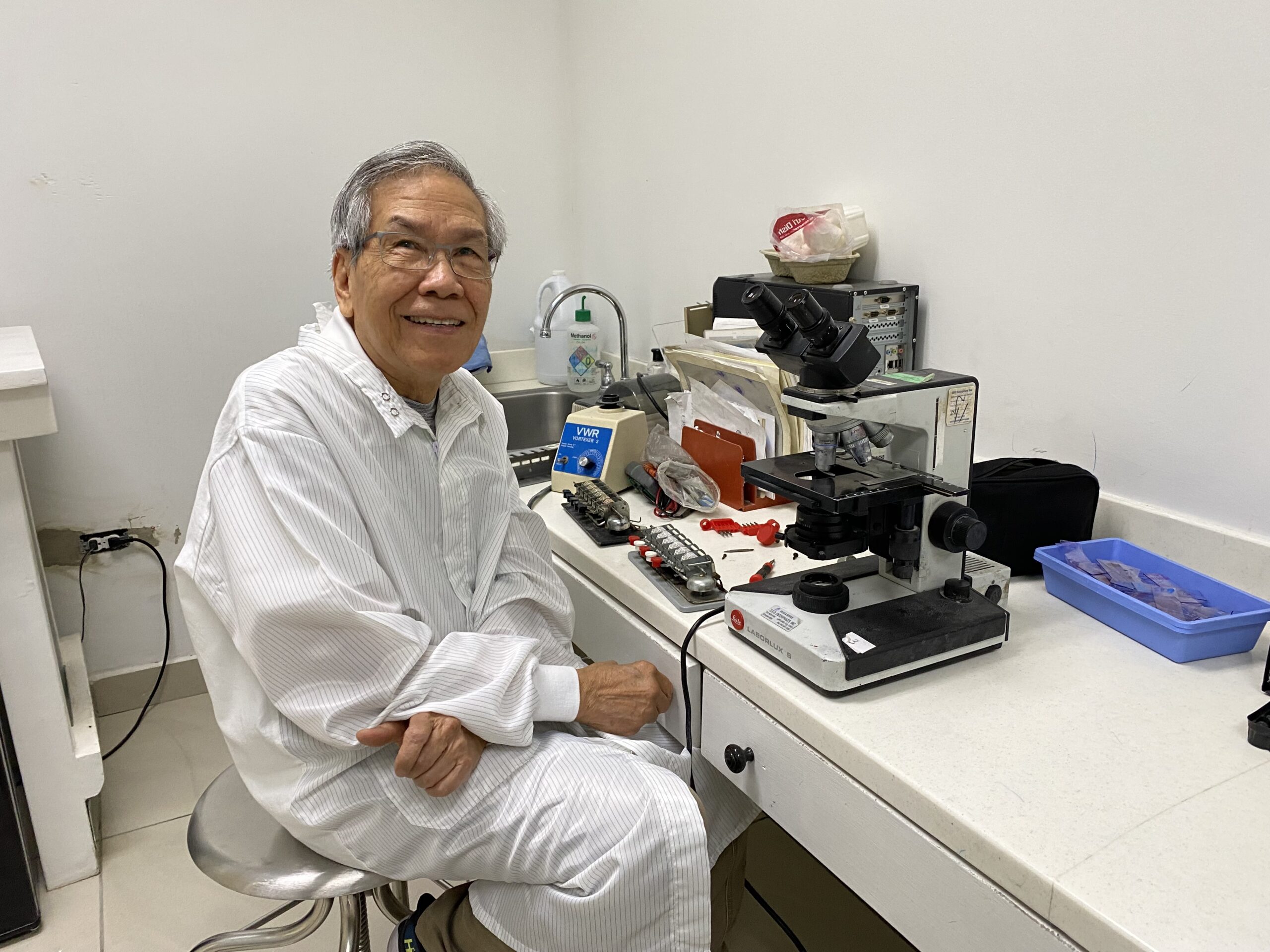 A man in a lab coat sitting next to a microscope. 